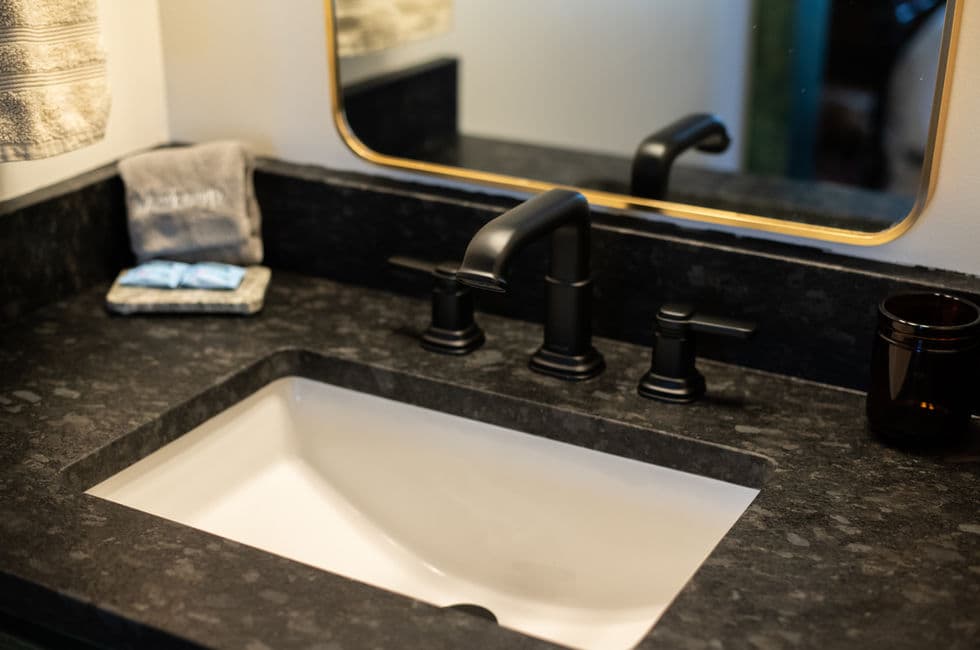 The bathroom sink with rectangular white basin, stone slab top, and black faucet and handles.