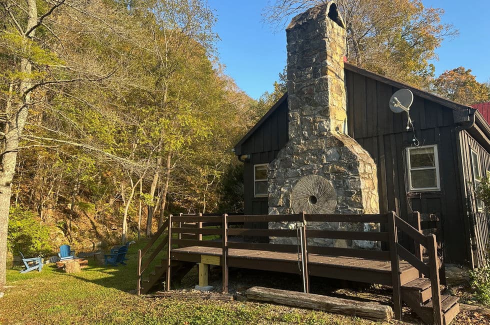 Exterior view of rustic wood framed cabin with large stone chimney fitted with an old mill stone and a wooden deck with stairs.