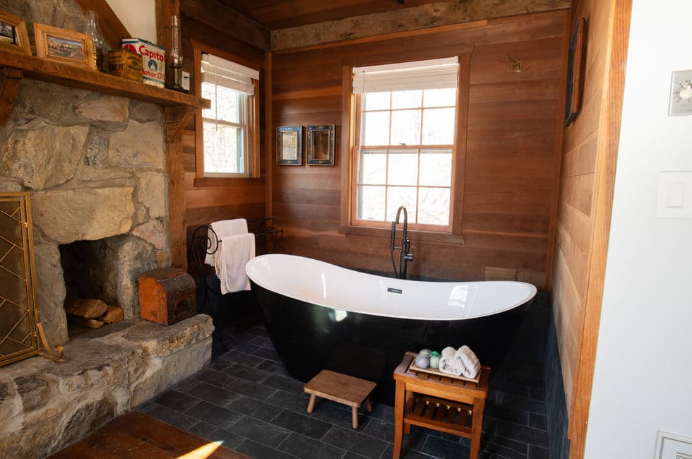 Rustic cabin interior with large stone fireplace, wood paneled walls, and black & white slipper tub.
