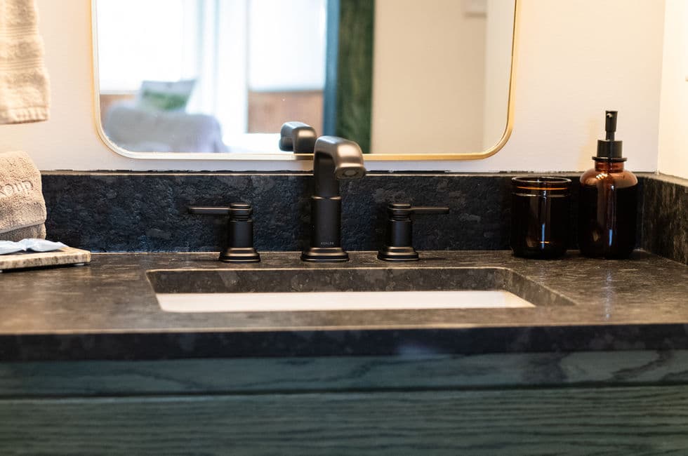 The bathroom sink with rectangular white basin, stone slab top, and black faucet and handles.