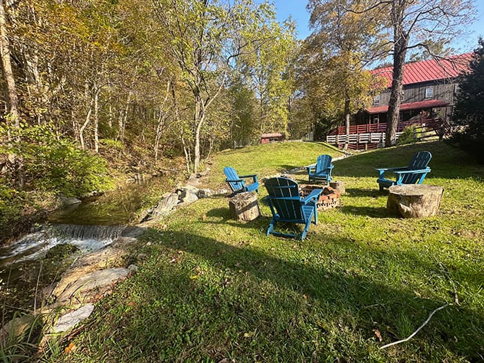 A scenic outdoor area features blue Adirondack chairs arranged around a fire pit beside a stream and a house in the background.
