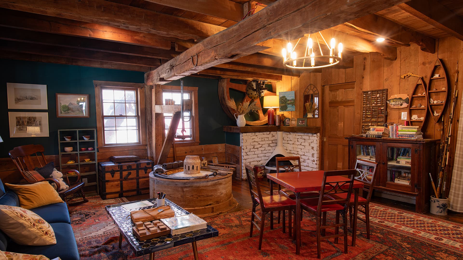 A cozy, rustic living room featuring wooden beams, a rocking chair, a dining table, and a decorative fireplace.