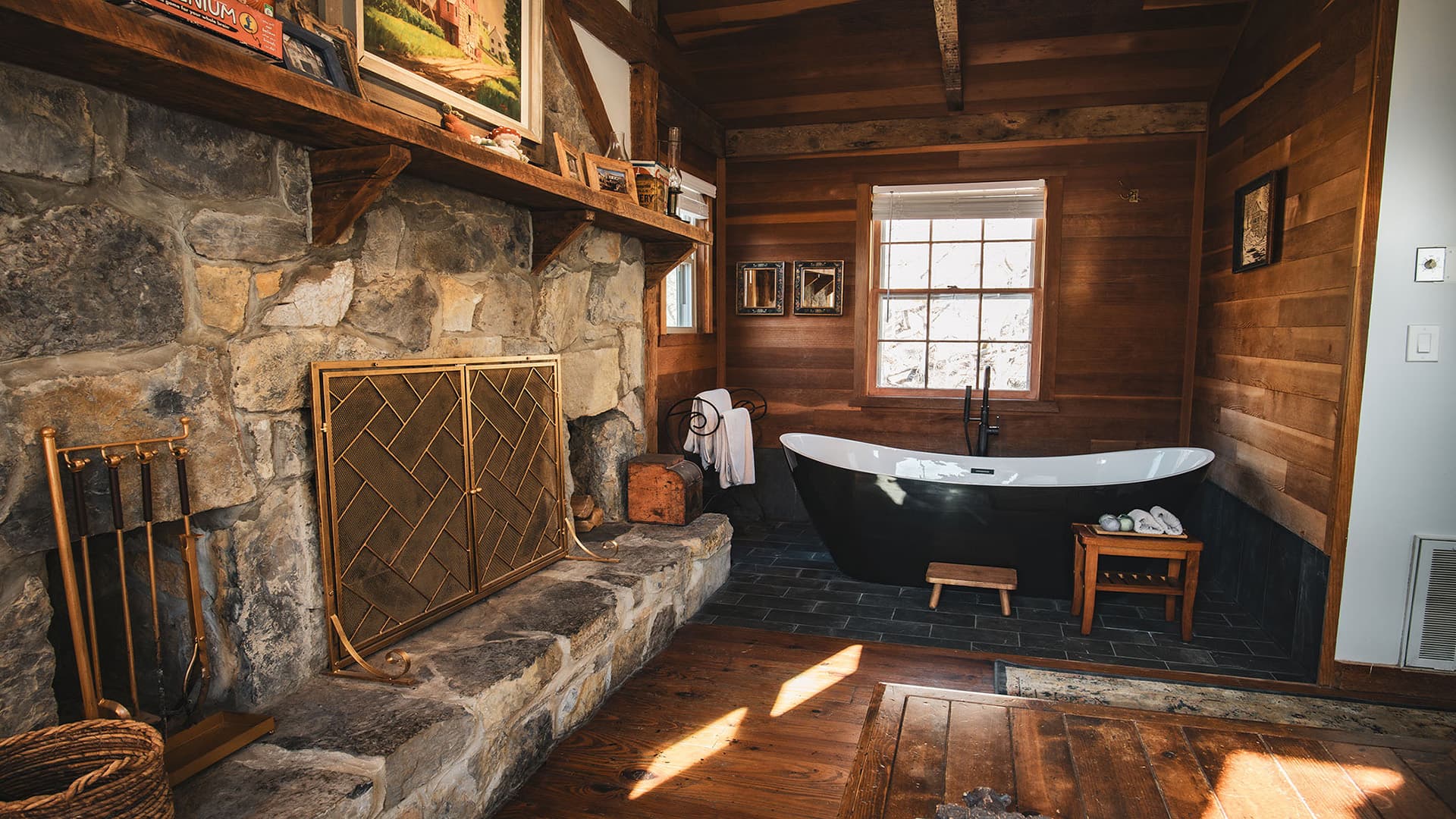 A rustic bathroom featuring a black freestanding bathtub, stone walls, and wooden accents.